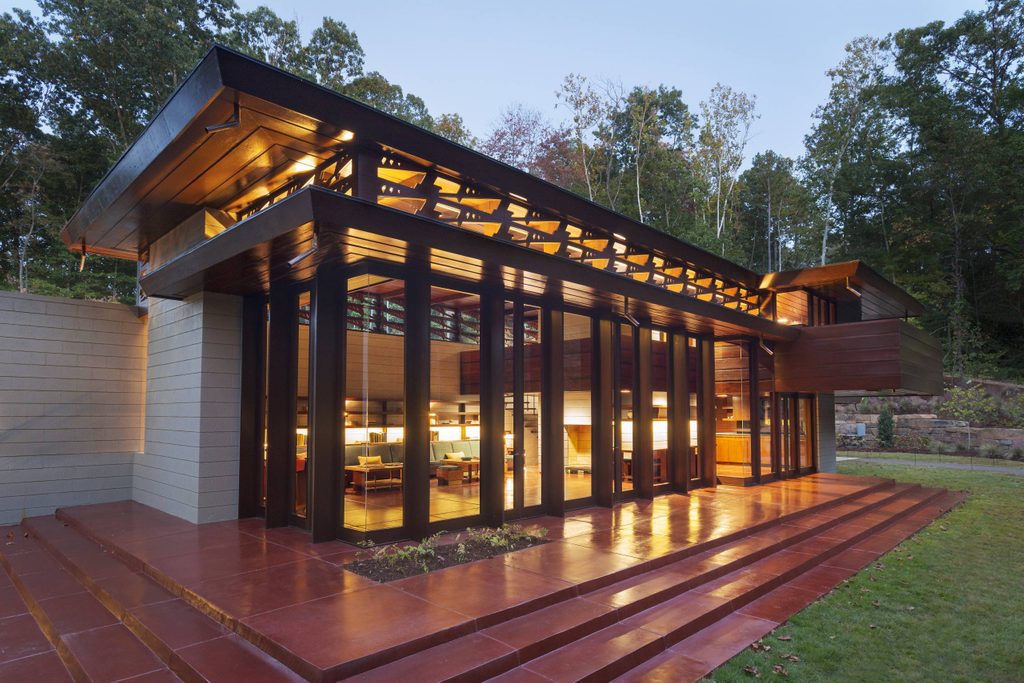 Modern glass and wood building with lit interior and terraced steps, set in a wooded landscape.