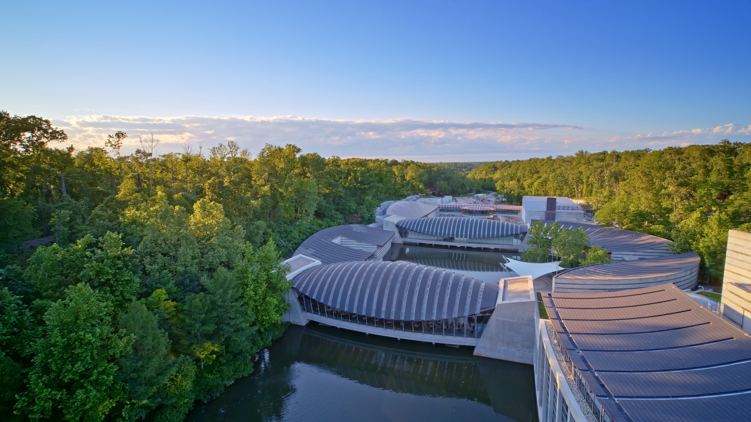 Modern architectural complex with curved ribbed roofs over water surrounded by dense green trees.