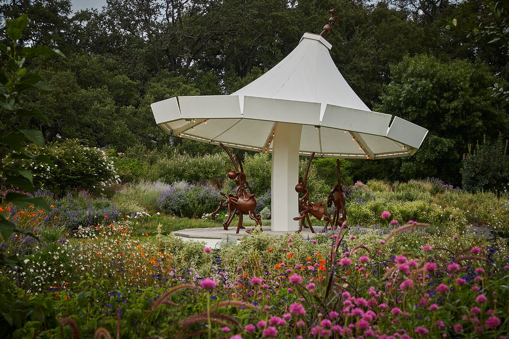 Carousel-like sculpture with white canopy and abstract brown horse figures in a flower garden.