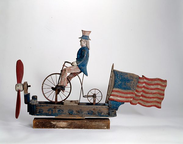 Wooden sculpture of a character in a blue coat and hat on a bike with flag and propeller.