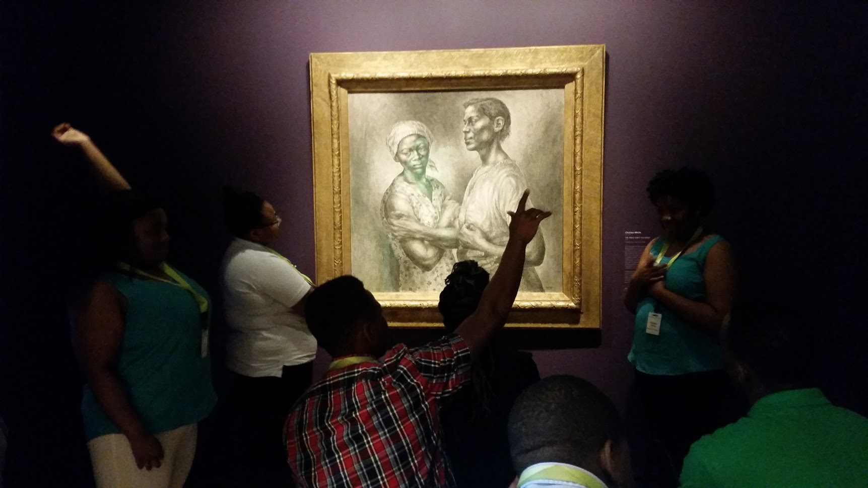 Group observing a framed artwork of two individuals in a dimly lit gallery, some pointing.