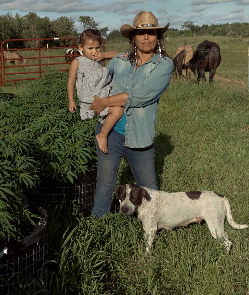 Photo of woman standing in field with cattle behind her holding a child and a dog standing at her knee