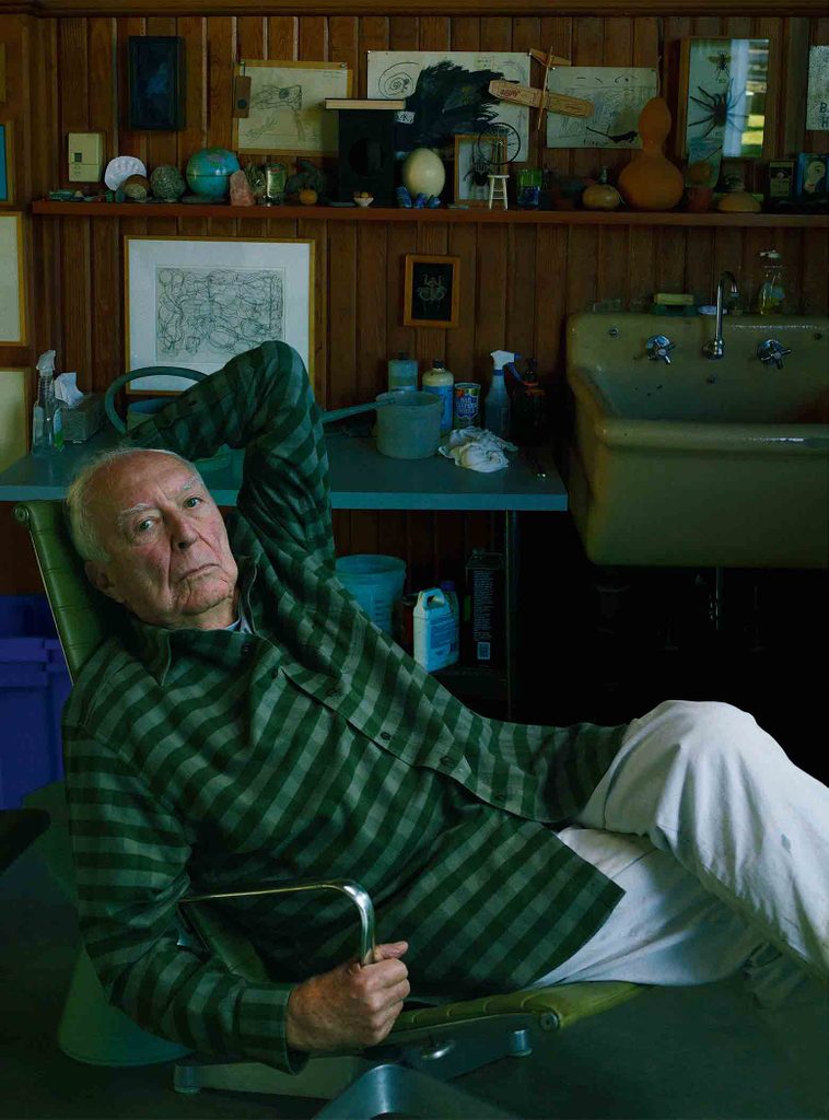 A photographed portrait of artist Jasper Johns, who looks at the camera as he reclines in a green chair in his painting studio.