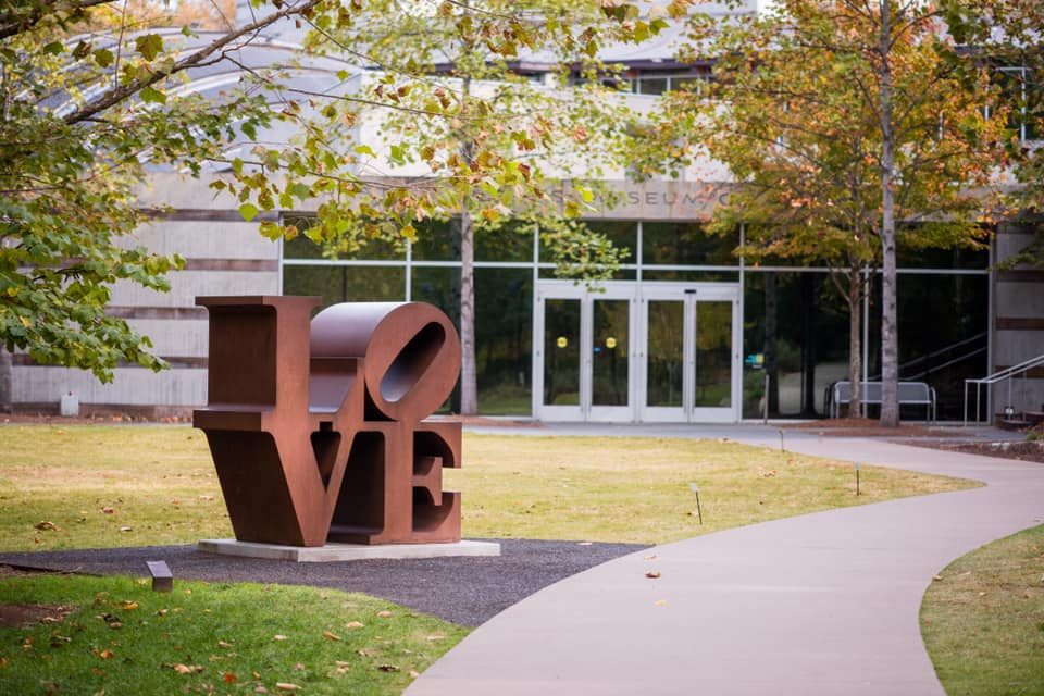 Rust-colored LOVE sculpture on grass in front of a modern building with autumn trees.