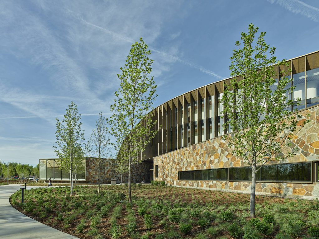 Modern building with stone facade and large windows surrounded by young trees and landscaping under blue sky
