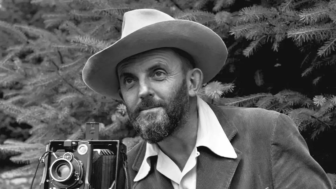 Man with beard in wide-brimmed hat, suit jacket, holding vintage camera, black and white photo.