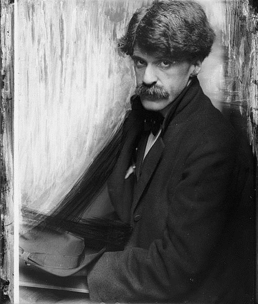 Man in suit with mustache holding hat, sitting by painted backdrop in aged black-and-white photo.