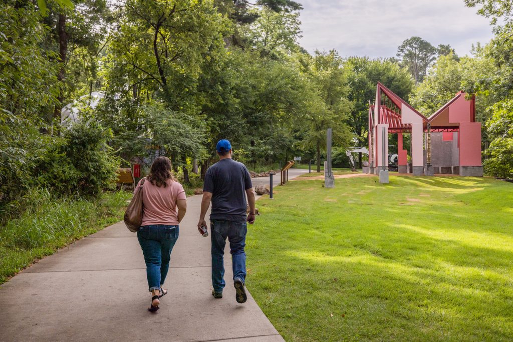 Two people walk along a paved trail lined with trees and grass. A small and angular pink house and other structures are visible among the trees ahead of them.