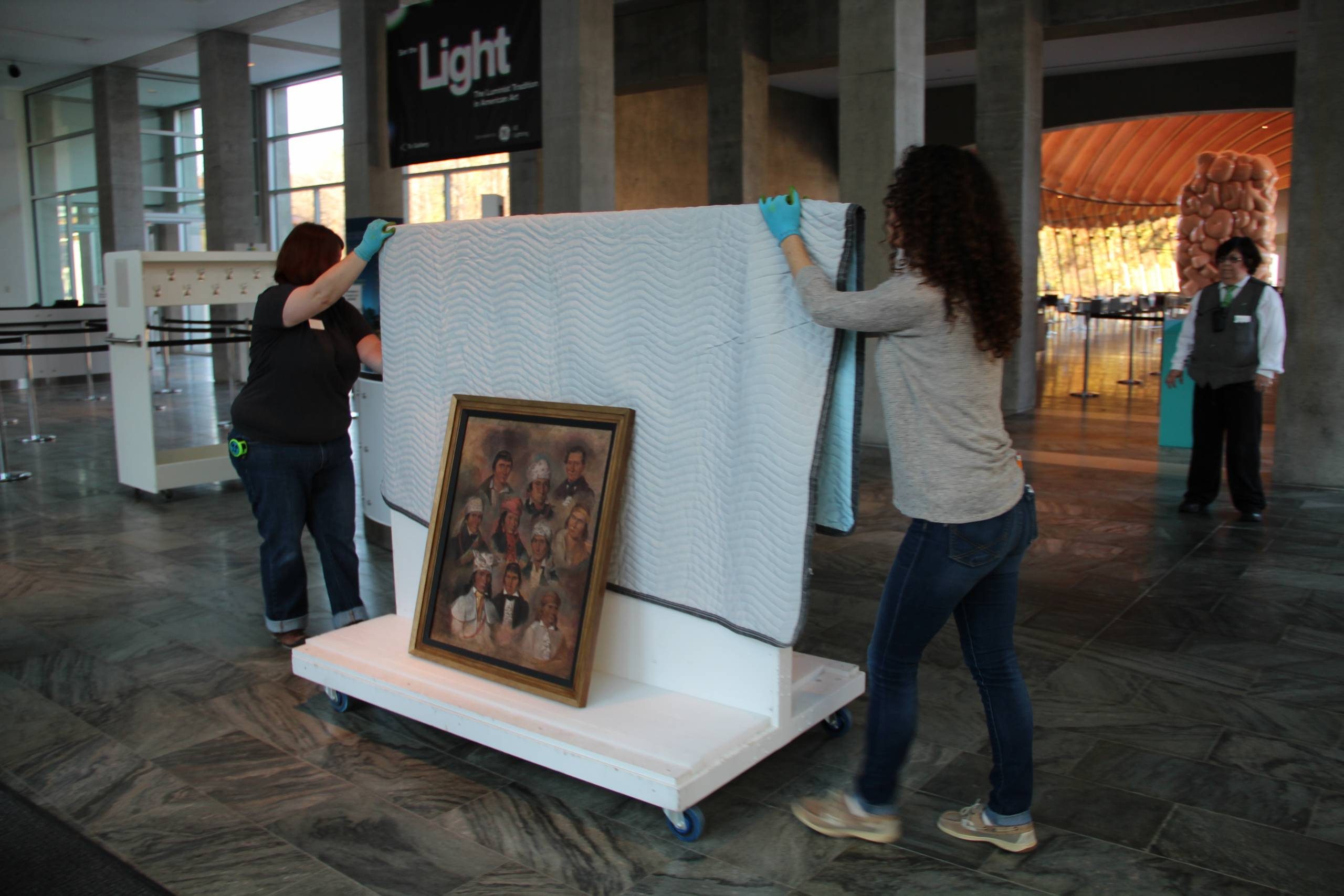 People moving a wrapped painting on a cart in a modern interior space, with an observer nearby.