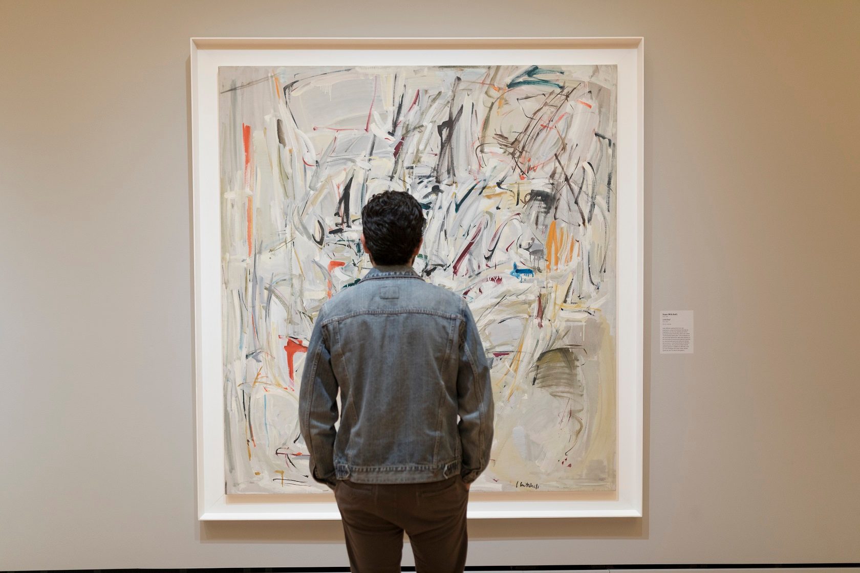 Person in denim jacket views colorful abstract painting in gallery with info plaque.