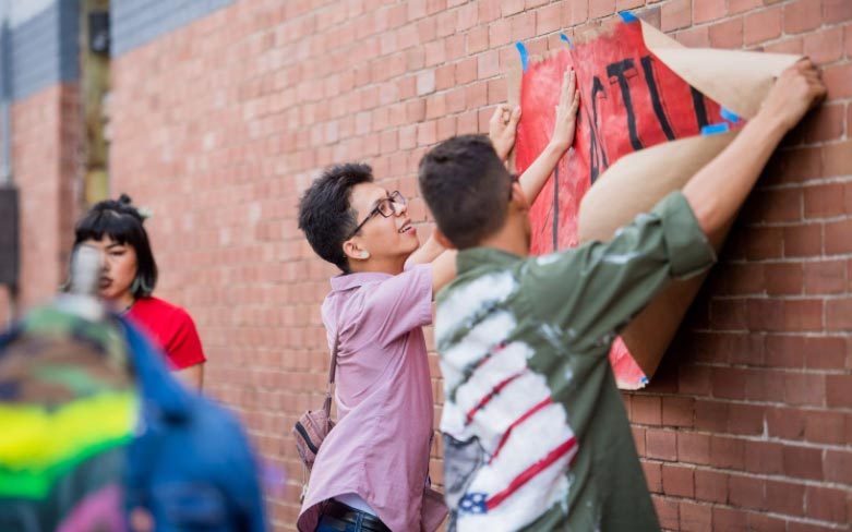 People hanging a large, red poster on a brick wall using tape.