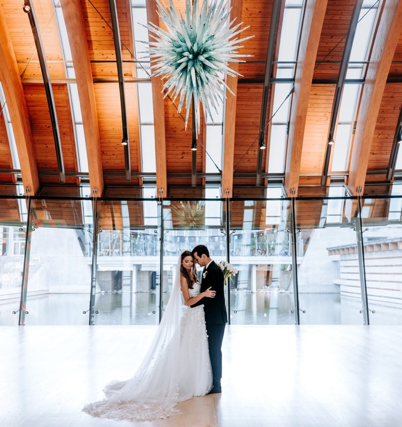 Bride and groom embrace in modern space with glass walls, wooden beams, and spiky chandelier.
