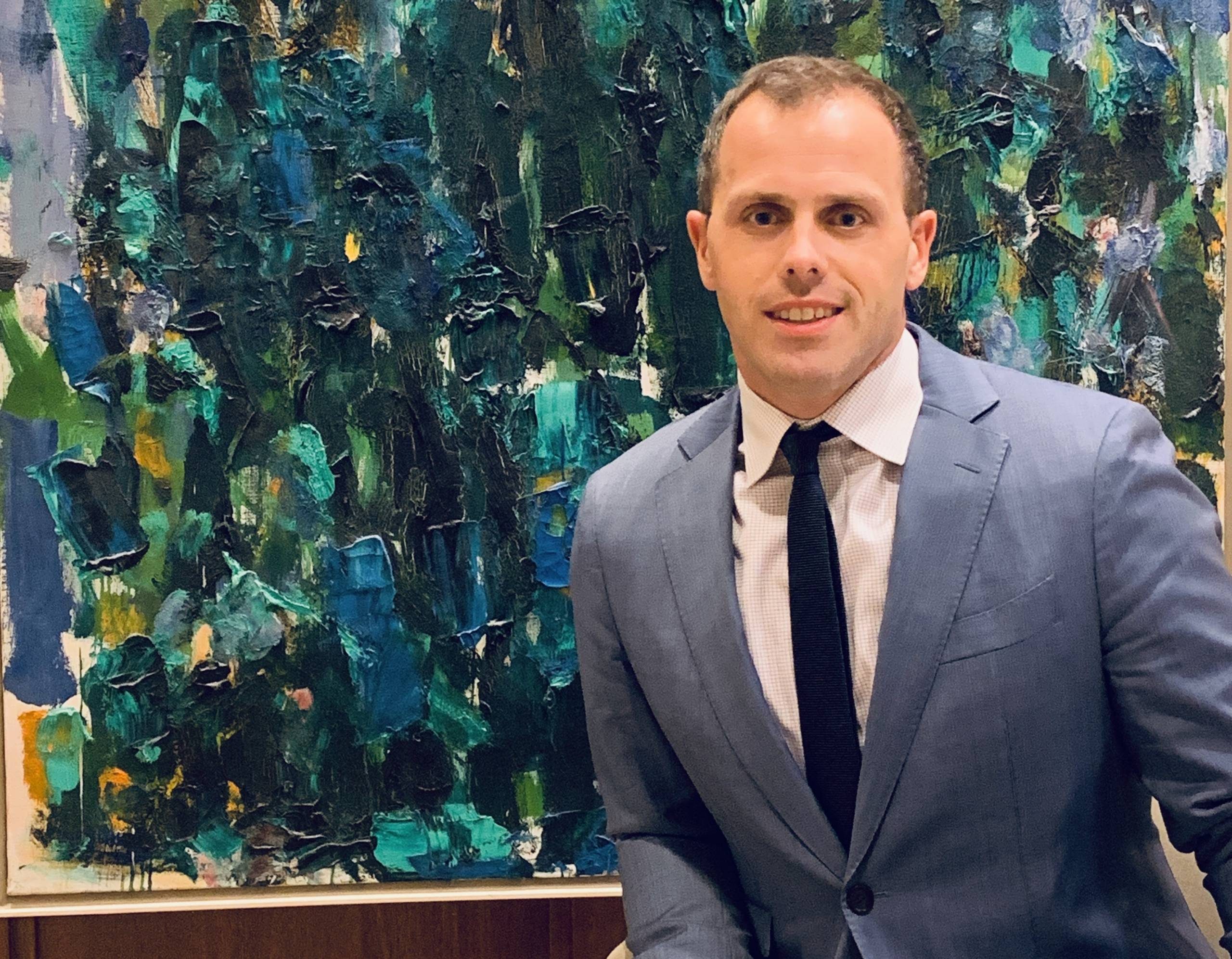 Person in a light gray suit with black tie in front of abstract blue and green painting.