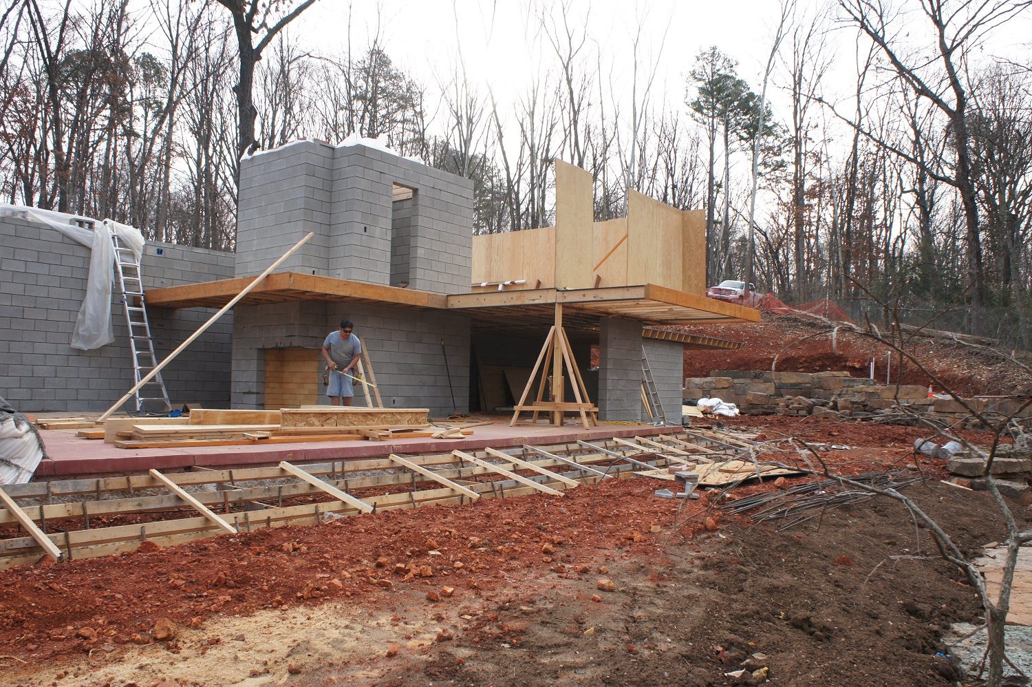 Construction site with concrete blocks, wooden beams, worker, materials, ladder, trees, red soil.