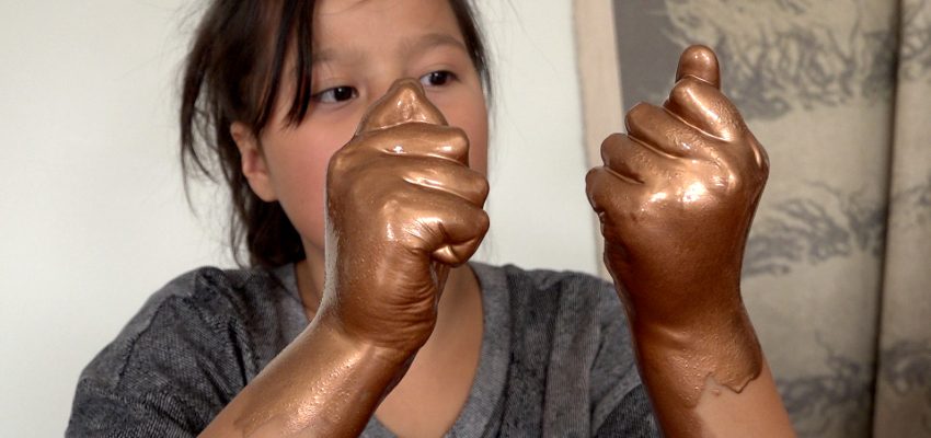 Child with bronze-painted hands, hair tied back, patterned curtain in background.