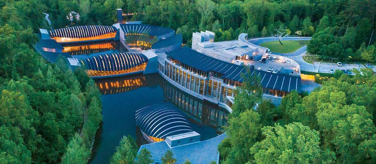 Aerial view of a modern complex with curved roofs, surrounded by lush forest and water.