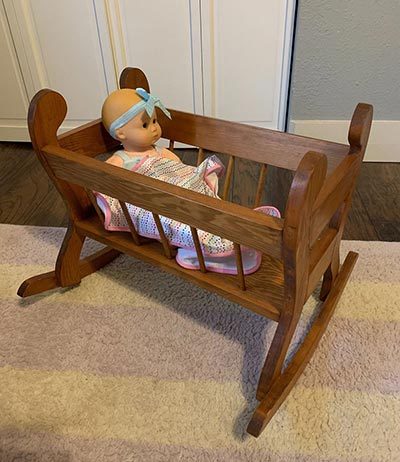 Wooden rocking cradle with a baby doll in a white outfit and blue headband on a patterned rug.