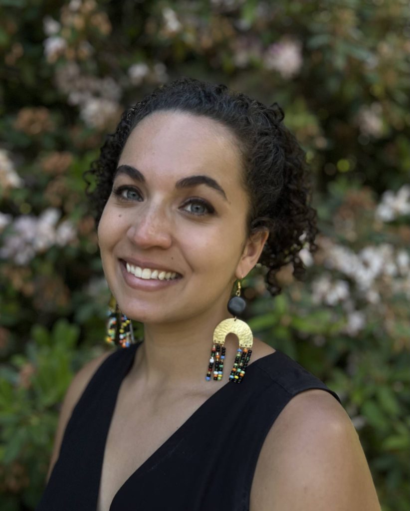 Woman with curly hair, smiling outdoors, wearing beaded earrings and black sleeveless top.