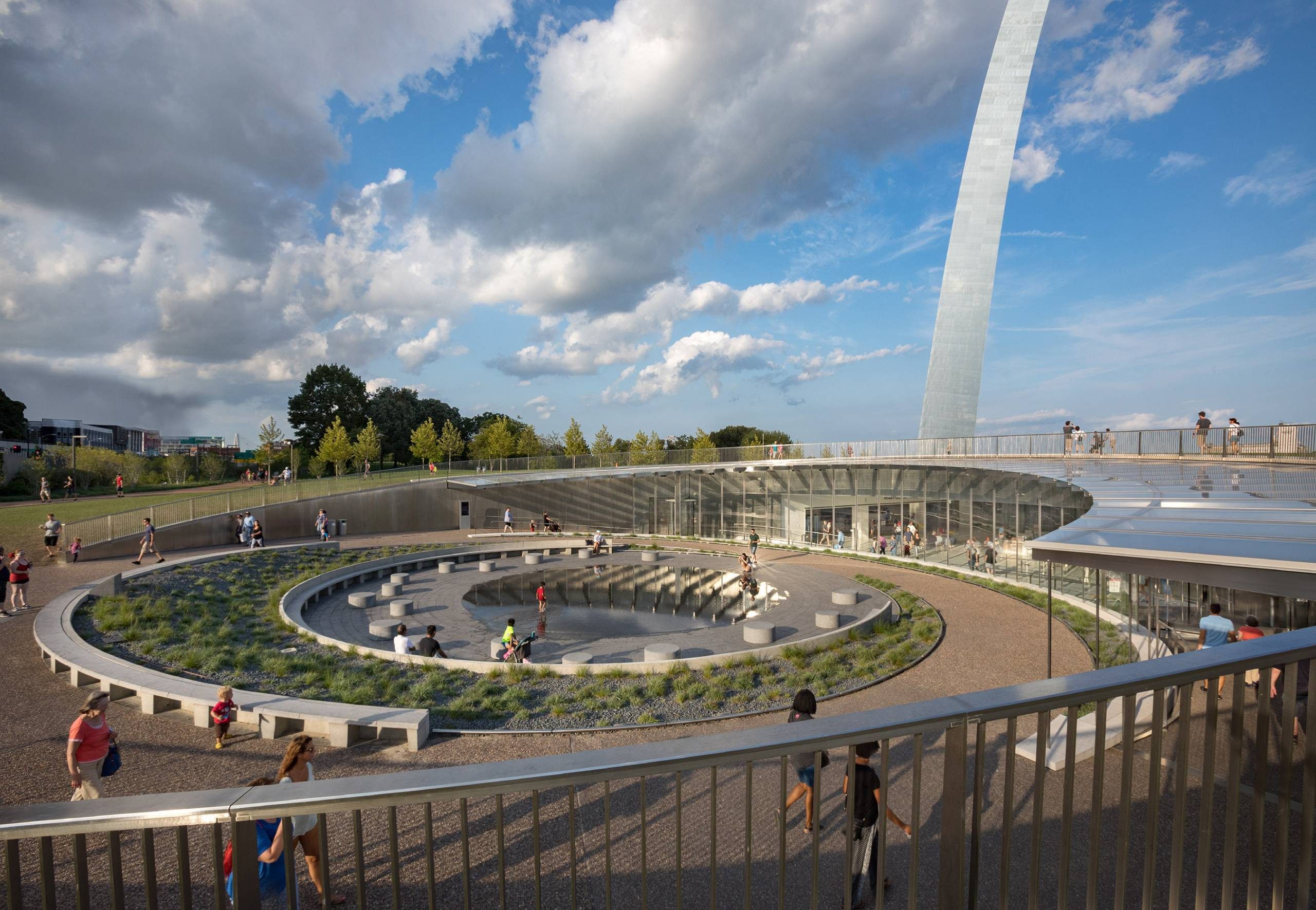 Modern outdoor space with circular seating, landscaping, people, pool, and upward arch structure.