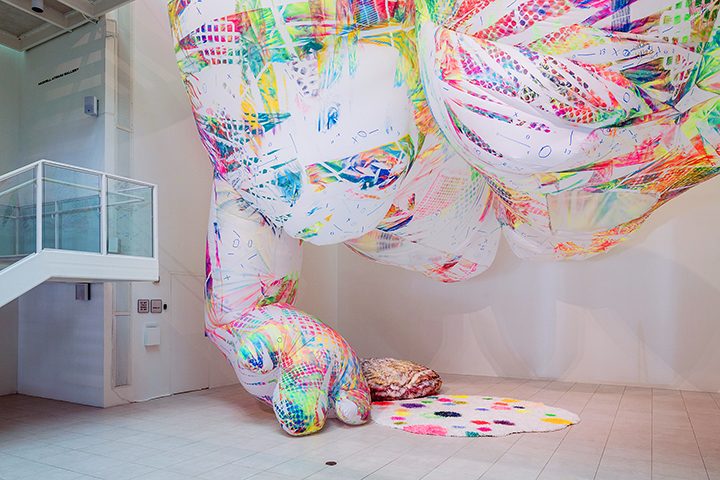 Colorful abstract fabric installation with patterns, hanging from gallery ceiling above mats.
