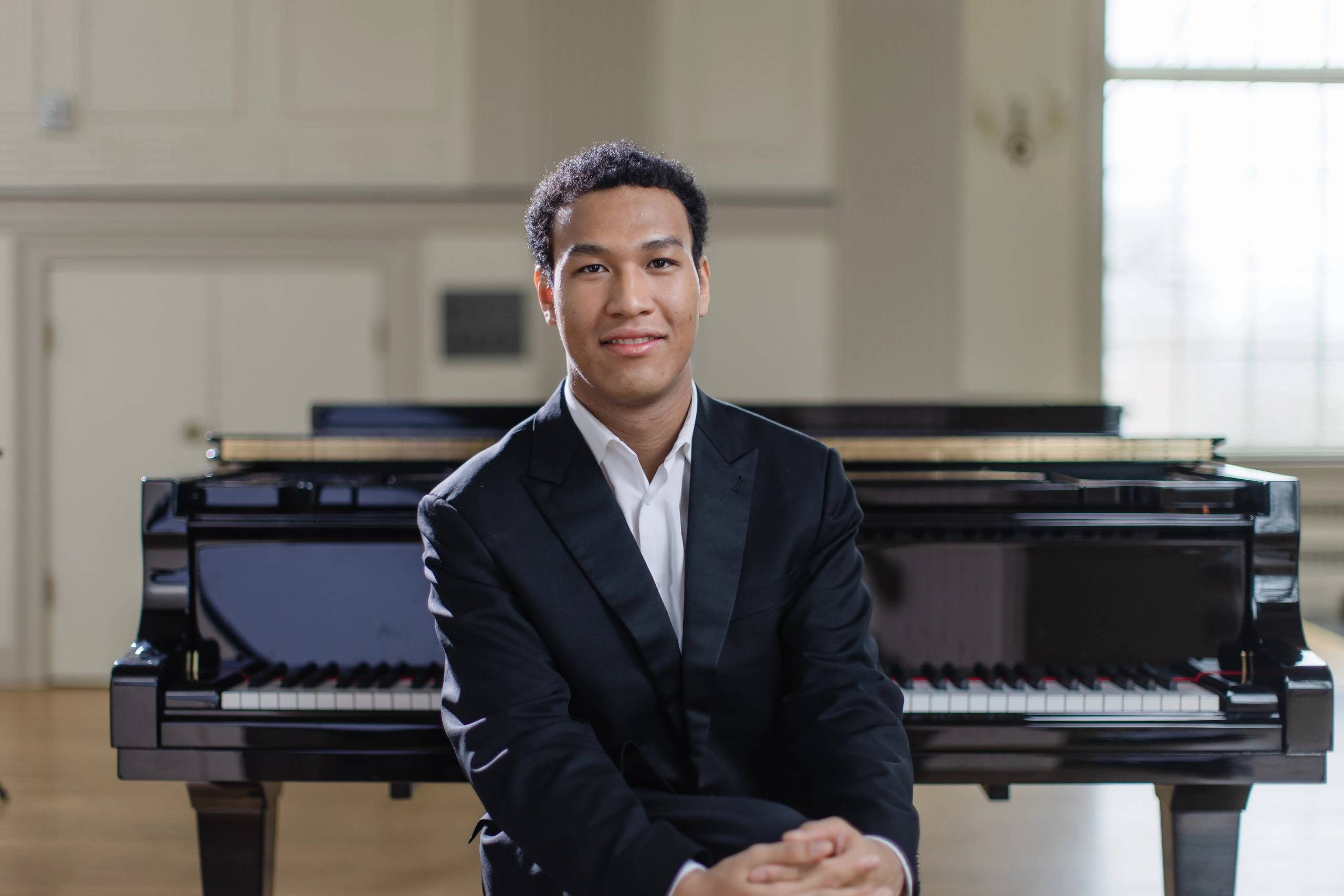 Person in a suit seated by a grand piano in a bright room.