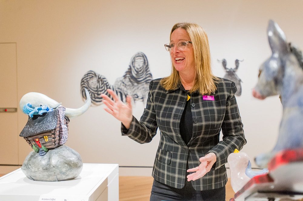 Woman in checkered blazer speaks near a house sculpture with tail in an art gallery.
