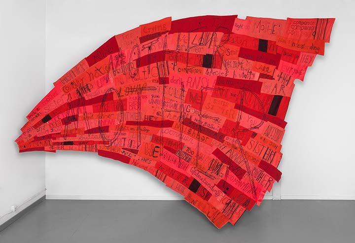 Wall-mounted art with red and orange fabric panels, handwritten text, abstract shape, white wall.