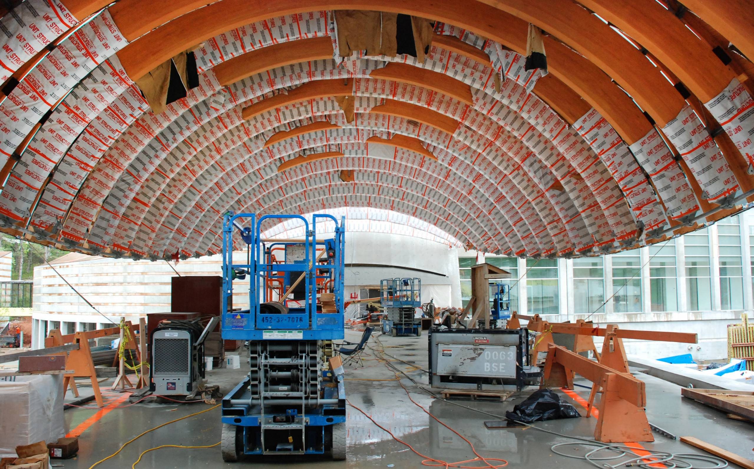 Construction site with curved wooden ceiling, blue lift, tools, and glass windows in background.