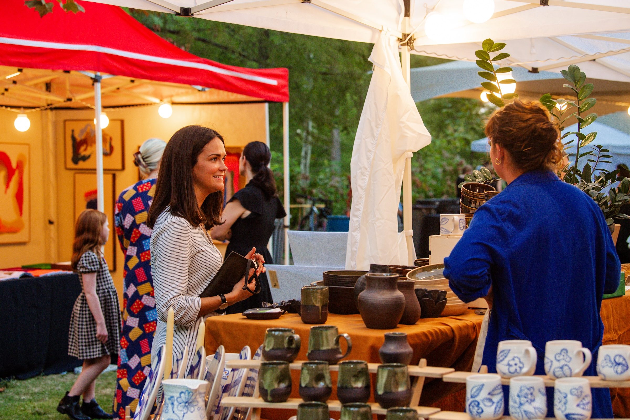 Two women converse at an outdoor market stall with ceramic pottery under tents.