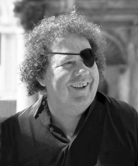 Curly-haired person with eyepatch and dark shirt smiling outdoors, urban background blurred.