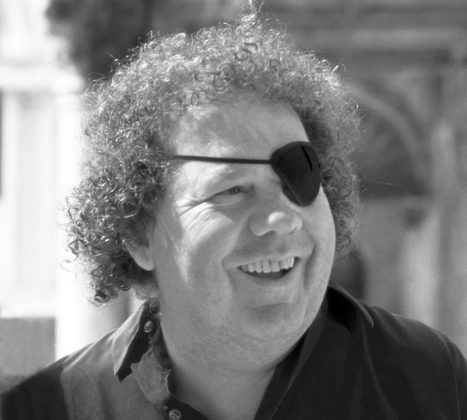 Person with curly hair, wearing eye patch and collared shirt, smiling outdoors in black and white.