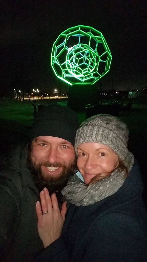 two guests pose in front of buckyball with an engagement ring