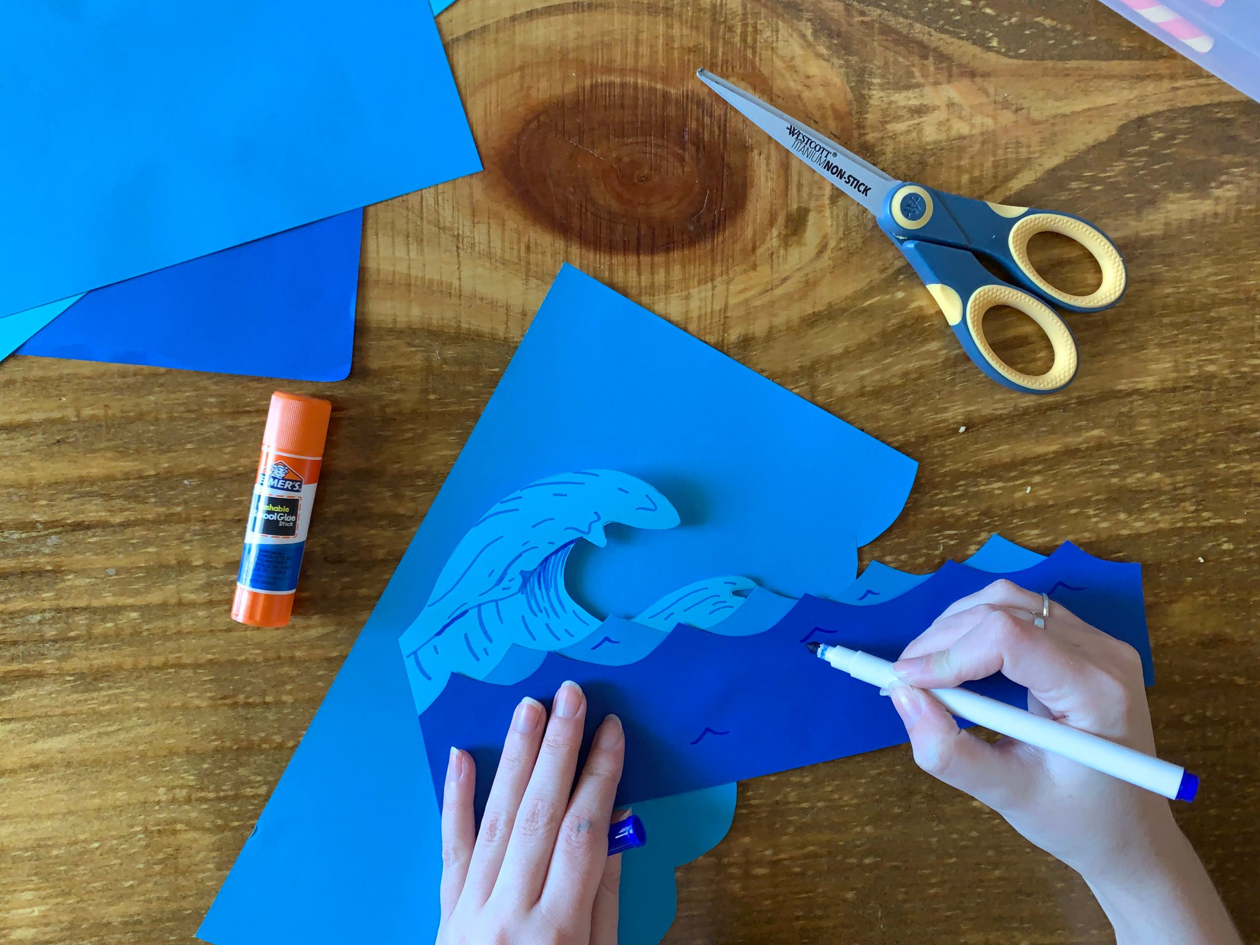 Crafting waves with blue paper, scissors, glue, and marker on a wooden table.