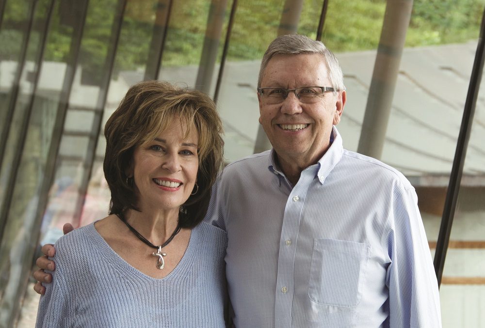 Older couple smiling indoors with large windows and greenery in the background.