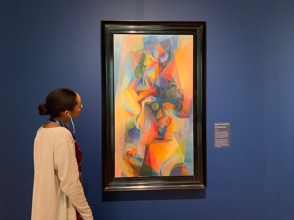 Una mujer de pie con audífonos en sus orejas observando una obra de arte que cuelga en una pared