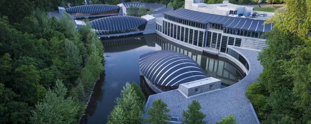 Modern architectural complex with curved roofs amid greenery, encircling a reflective water body.