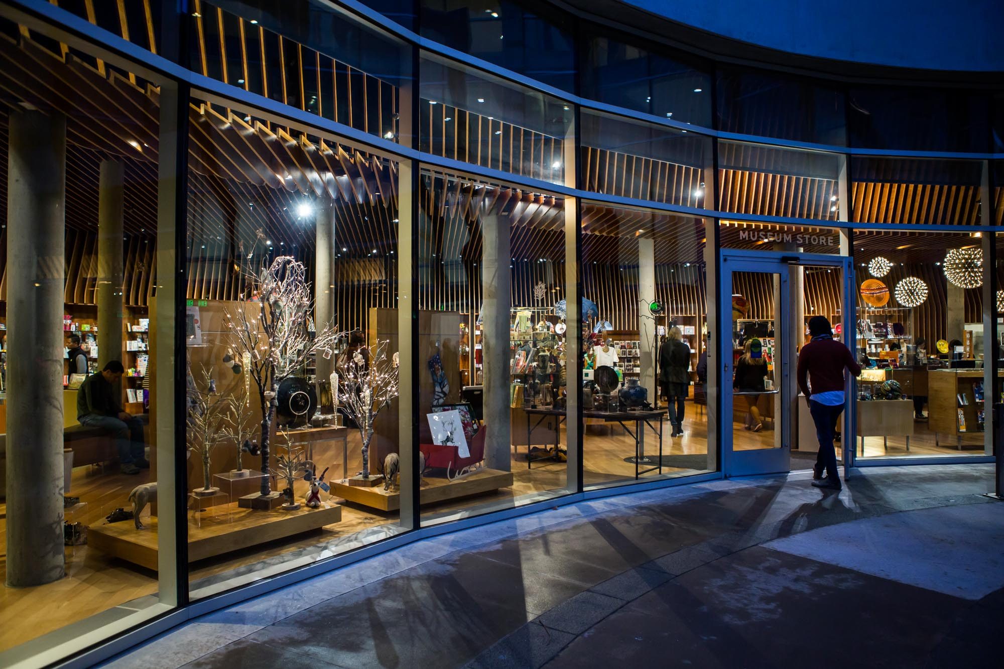 Museum store entrance with glass doors, modern design, sculptures, souvenirs, well-lit interior.