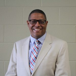 Person in glasses, beige suit, multicolored tie smiling in front of light brick wall.