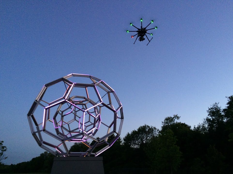 Geometric sculpture outdoors with drone above, trees and evening sky in background.