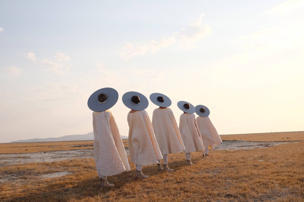 Five women dressed in white dresses and hats and standing in a grassy field
