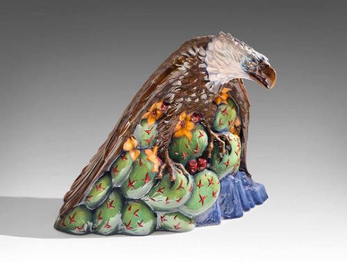 Ceramic sculpture of an eagle perched on green prickly pear cactus with yellow flowers on a blue base