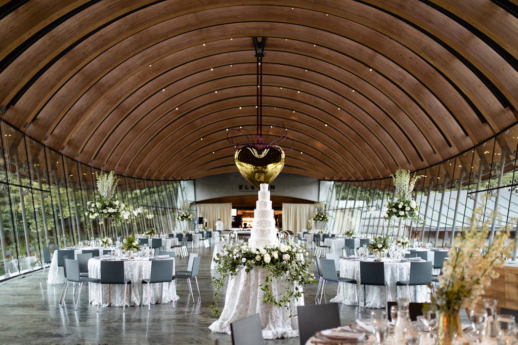Elegant event venue with arched ceiling, glass windows, formal tables, floral centerpieces, cake.