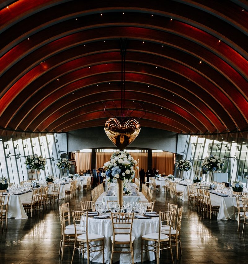 Reception hall with round tables, white tablecloths, gold chairs, floral centerpieces, large windows.