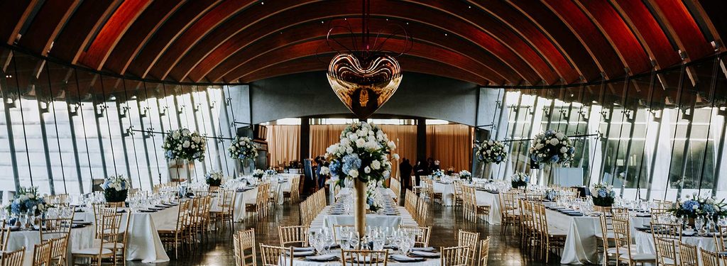 Banquet hall with vaulted wooden ceiling, glass walls, floral centerpieces, and gold chairs.