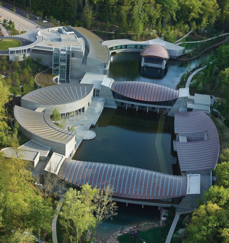 Aerial view of curved architectural complex around a pond, surrounded by greenery and walkways.