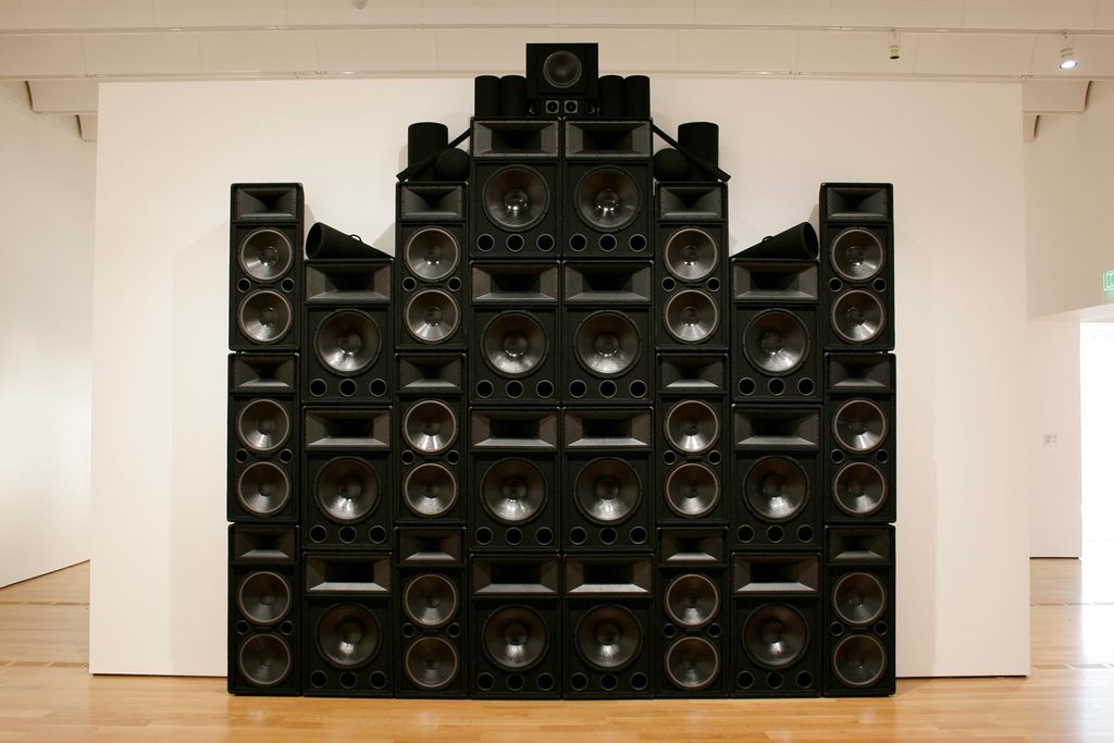 A large structure of stacked black speakers, black megaphones and black cylinders towers next to a white wall