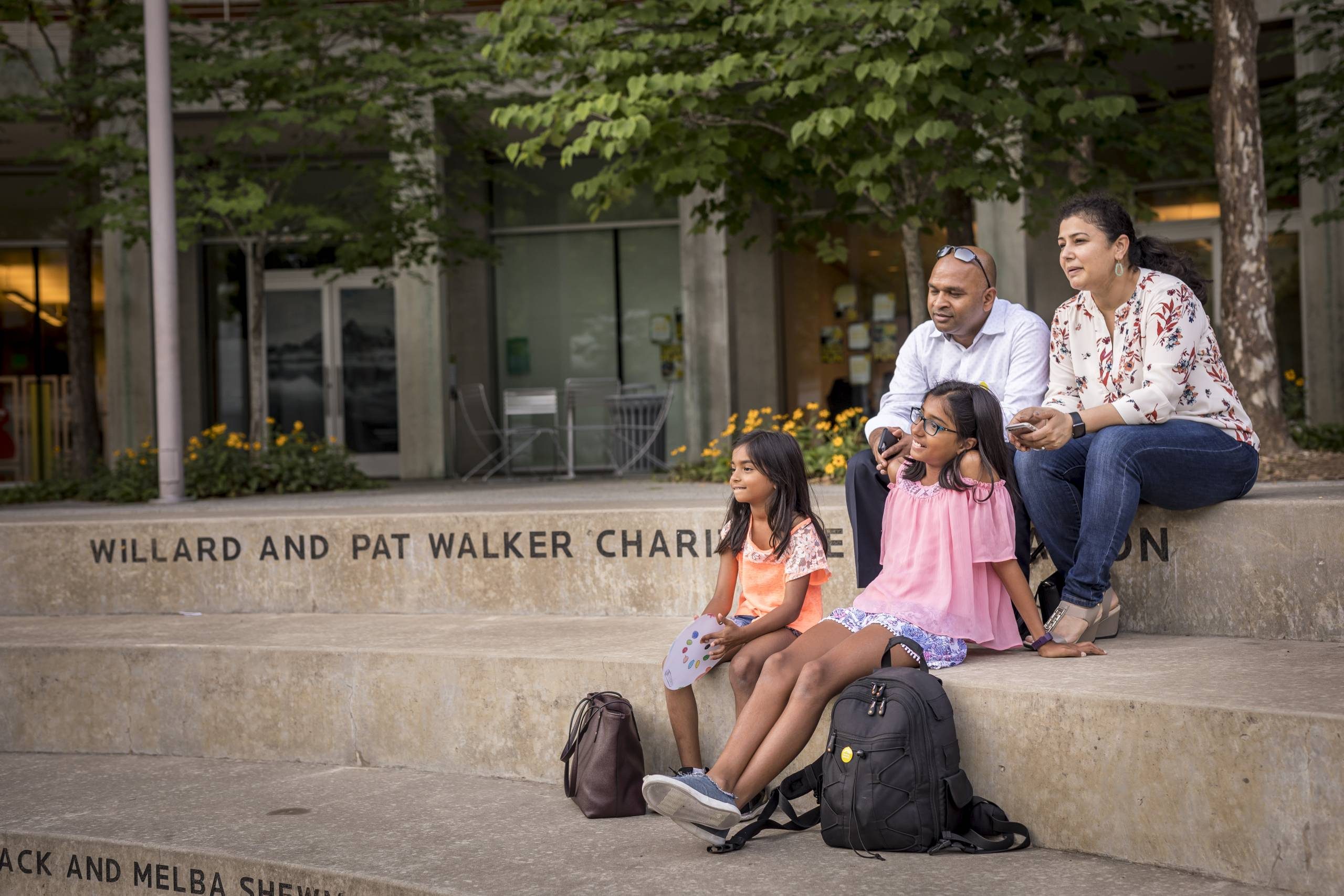 Family of four seated on steps outdoors, with backpacks, near a glass-windowed building.