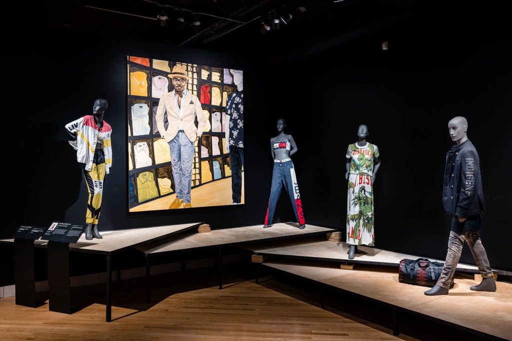 a gallery view centered on a large painting of a nicely dressed man with mannequins showcasing fashion garments on either side