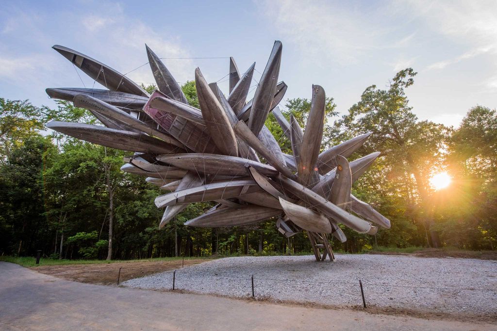 Outdoor sculpture of numerous metallic canoes suspended in a dynamic arrangement amidst trees.