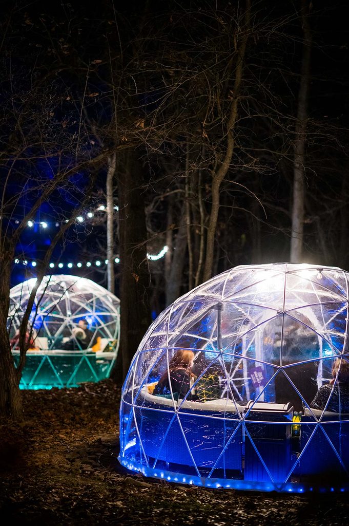 Illuminated geodesic domes in wooded area at night with outdoor dining inside.